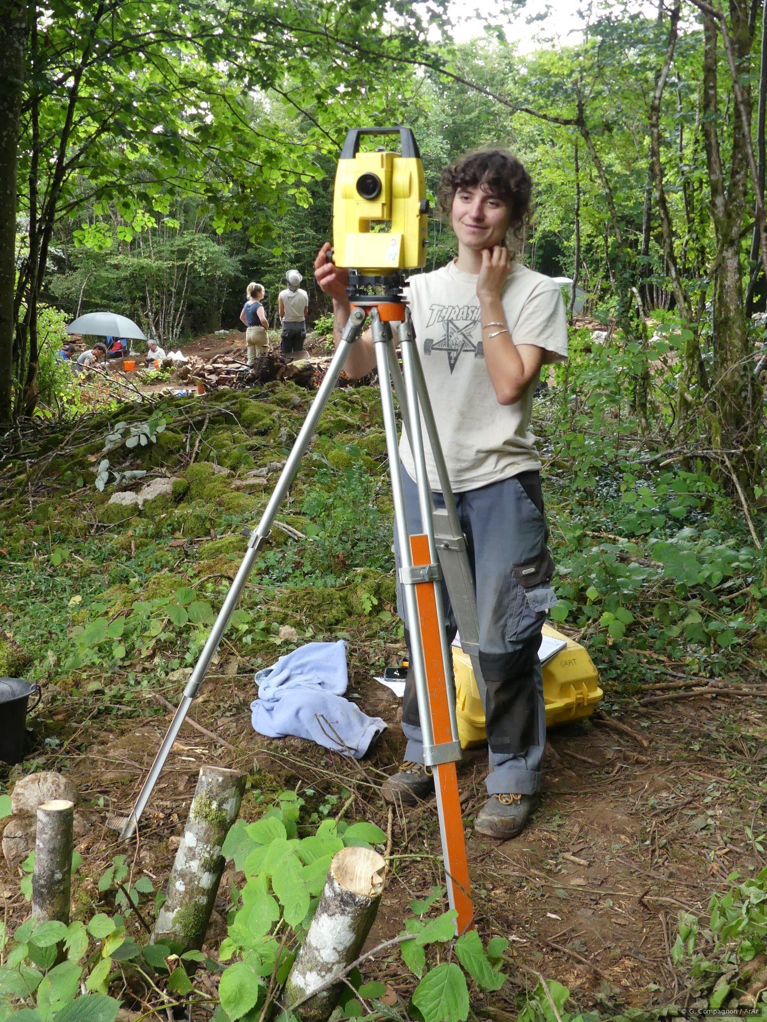 Étudiante manipulant un tachéomètre laser sur le chantier de fouille, en forêt.